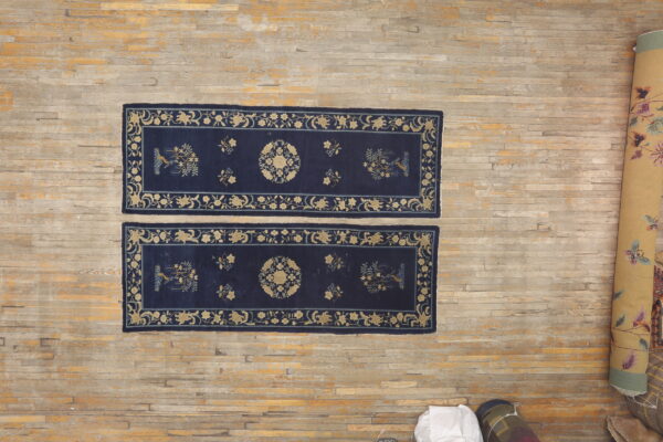A pair of navy and gold patterned runners lie on a wood floor beside several rolled rugs.