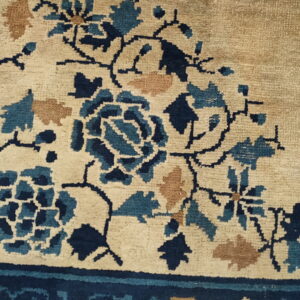 Close-up of a rug showing beige pile with navy blue, teal, and tan stylized floral patterns near a dark border.