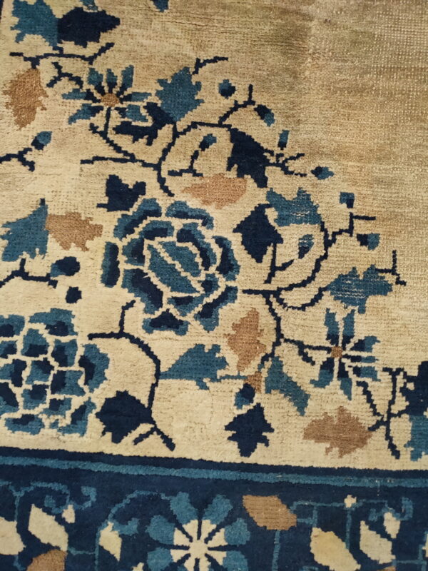 Close-up of a rug showing beige pile with navy blue, teal, and tan stylized floral patterns near a dark border.