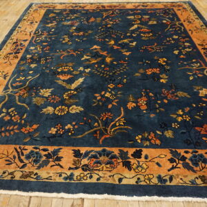 Navy floral rug with thick orange border lies on aged light hardwood flooring.