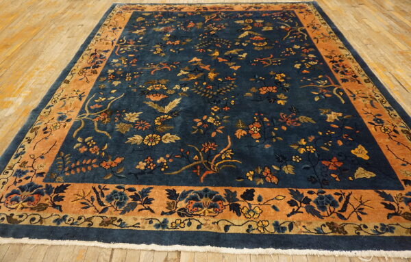 Navy floral rug with thick orange border lies on aged light hardwood flooring.