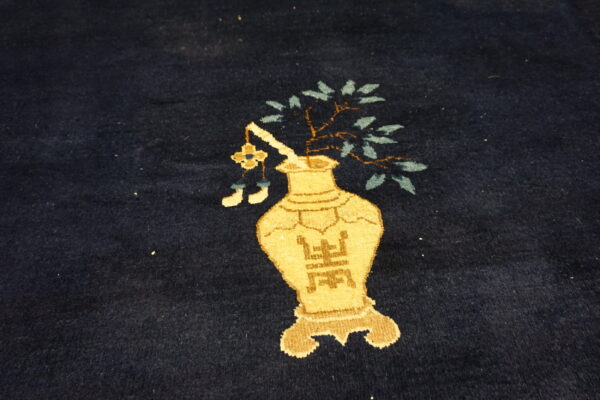 Close-up of dark blue pile rug featuring a tan vase design with stylized symbols and green leaves.