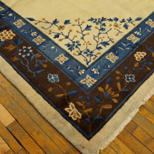 A rug corner with a cream field and dark brown and blue floral borders rests on wood flooring.