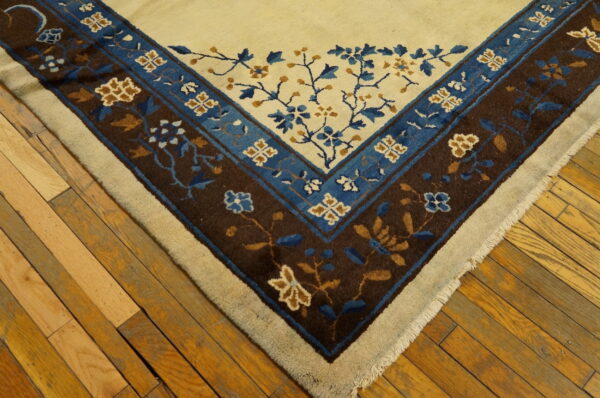 A rug corner with a cream field and dark brown and blue floral borders rests on wood flooring.
