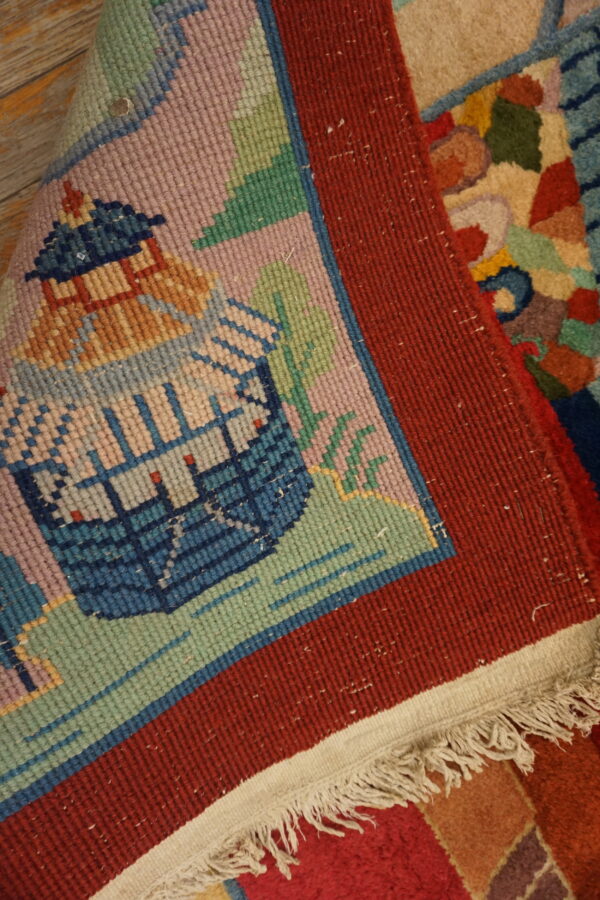 Detail of a low-pile rug showing a pink and blue pictorial field, wide red border, and white fringe.