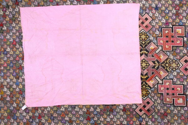 Pink quilted fabric covers a multicolored rug featuring dense geometric and cross-stitch patterns.