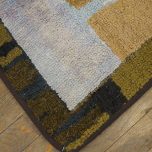 Chunky textured rug corner in beige, olive, light blue, and black geometric blocks rests on rustic wood flooring.