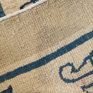 Textured beige rug with thick blue lines and abstract shapes is curled back showing the edge.