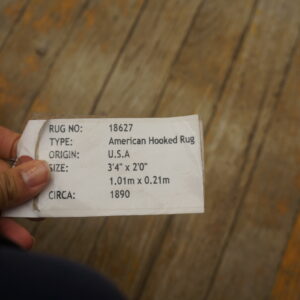 A hand holds a white descriptive rug tag above rustic brown wooden flooring with orange paint details.