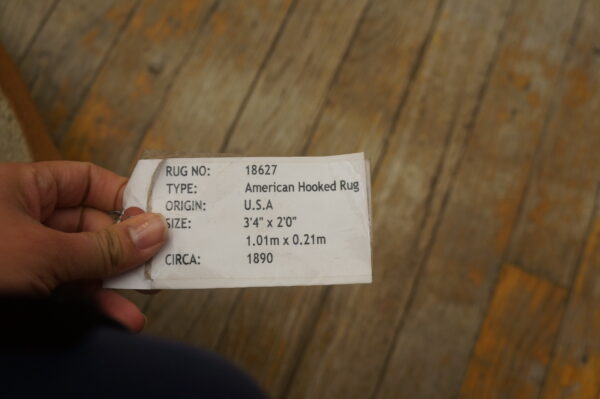 A hand holds a white descriptive rug tag above rustic brown wooden flooring with orange paint details.