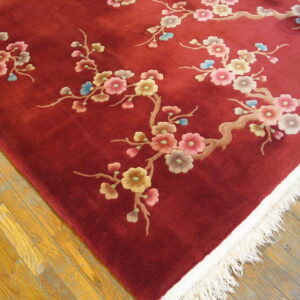 Close-up of a high-pile burgundy rug with floral branches and cream fringe on golden wood flooring.