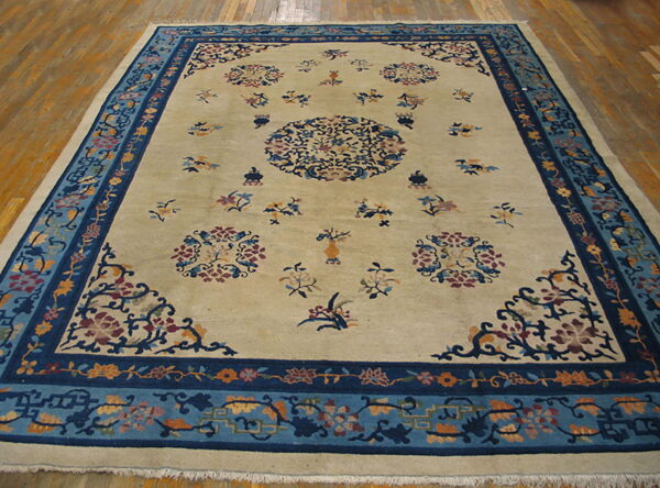 Large rectangular beige rug featuring intricate blue borders and scattered multicolor floral medallions on a wood floor.