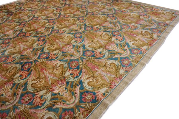 A detailed, high-pile rug featuring scrolling floral patterns in gold, teal, pink, and tan colors on a white background.