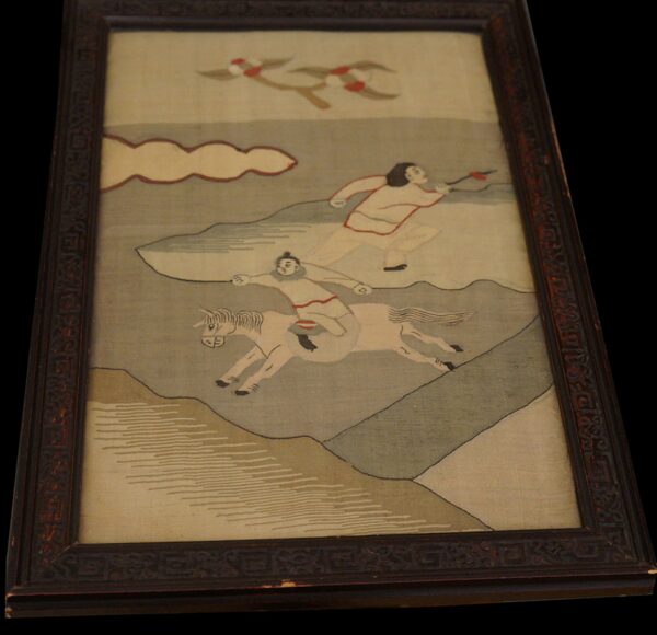 A framed textile displaying pale gray and tan figures, a horse, clouds, and birds against a neutral field.