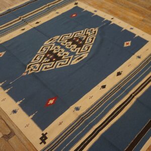 Blue flatweave rug with bold tan and black geometric patterns resting on worn hardwood flooring.