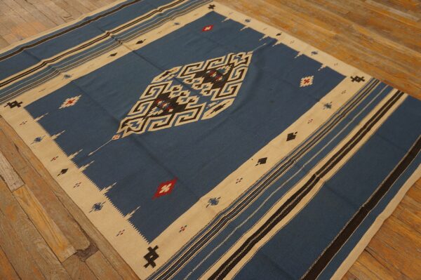 Blue flatweave rug with bold tan and black geometric patterns resting on worn hardwood flooring.