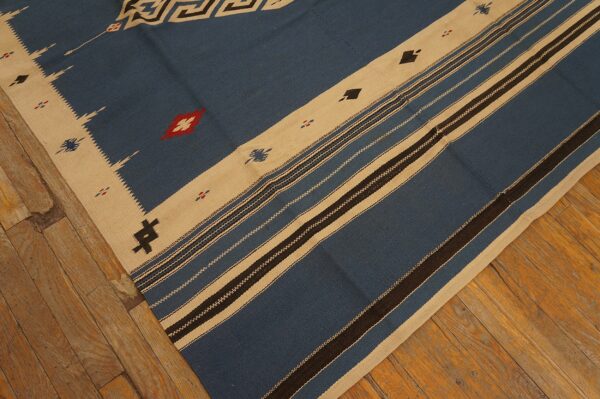 Blue flatweave rug with beige geometric borders and dark stripes laid on worn wood flooring.