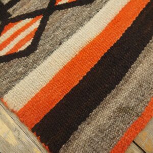 Geometric patterns and wide orange, gray, and brown stripes characterize this coarse woven rug on rustic wood.