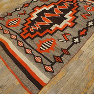 Gray flatweave rug with vivid orange, dark brown, and white geometric patterns on worn wood planks.