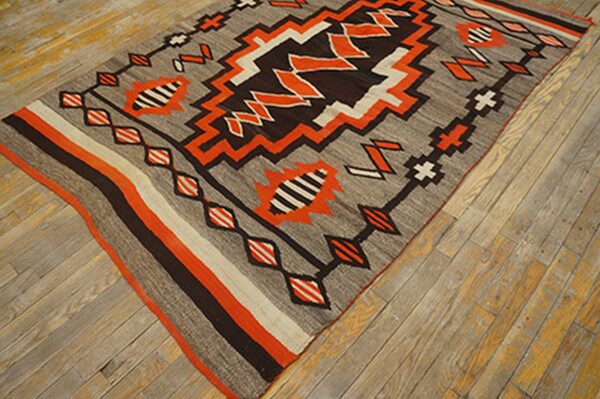 Gray flatweave rug with vivid orange, dark brown, and white geometric patterns on worn wood planks.