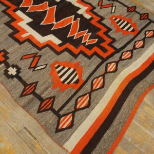 Flatweave rug with zigzag geometric patterns in orange, gray, dark brown, and cream on distressed wood flooring.