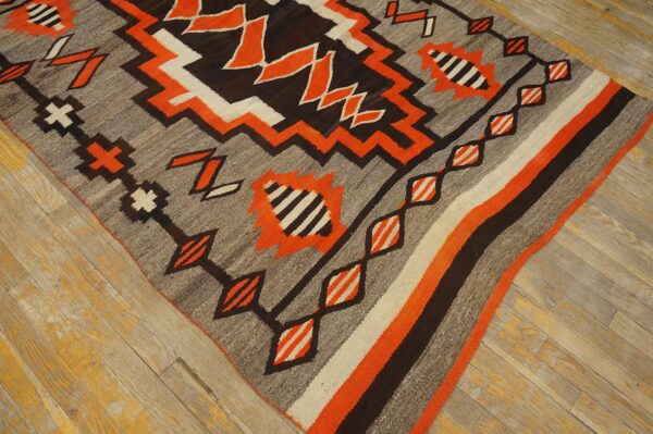 Flatweave rug with zigzag geometric patterns in orange, gray, dark brown, and cream on distressed wood flooring.