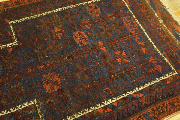 Worn, low-pile rug displaying intricate geometric patterns in navy, rust, and brown tones.