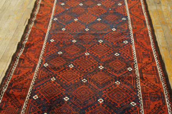 A red and dark blue geometric runner rug with diamond patterns and small white accents rests on light wooden floorboards.