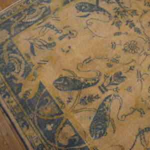 Faded low-pile rug in gold and indigo with stylized bird and floral patterns rests on wood planks.