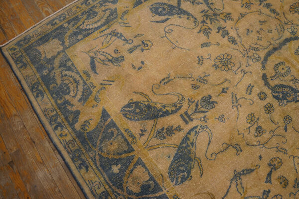 Faded low-pile rug in gold and indigo with stylized bird and floral patterns rests on wood planks.