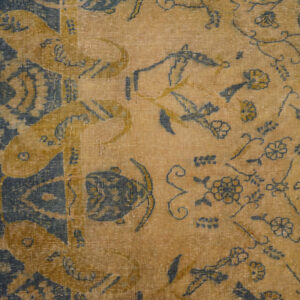 Distressed, low-pile rug detail in tan and dark blue with stylized floral patterns on wood flooring.