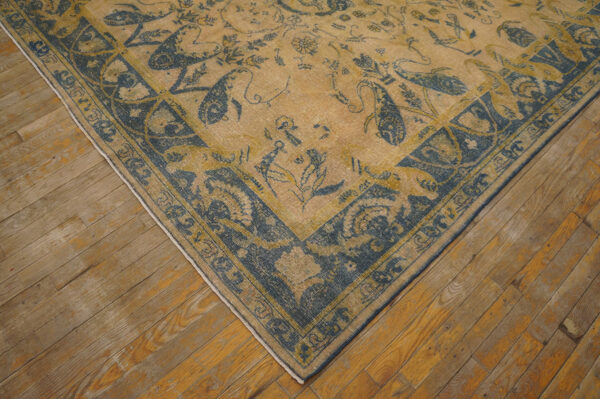 Low-pile beige rug with dark blue traditional motifs and borders sits on rustic wood plank flooring.