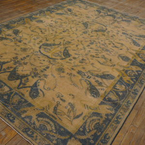 A tan rug with intricate indigo floral and bird motifs and a wide decorative border lies on wood plank flooring.