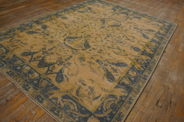 A tan rug with intricate indigo floral and bird motifs and a wide decorative border lies on wood plank flooring.