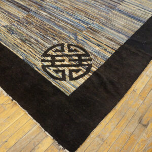 Striped blue and tan rug corner with a thick black border and geometric symbol lying on light hardwood flooring.