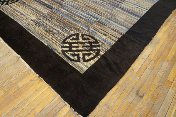 Striped blue and tan rug corner with a thick black border and geometric symbol lying on light hardwood flooring.