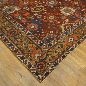 The corner of a traditional rug with a rust field and bold blue and gold border on light hardwood.
