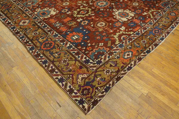 The corner of a traditional rug with a rust field and bold blue and gold border on light hardwood.