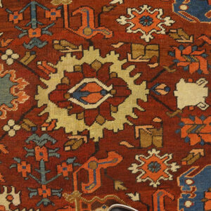 Close-up of a traditional rug with a deep rust background and geometric motifs in blue, gold, and cream.