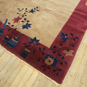 Tan field rug with a wide burgundy border featuring blue flowers and vase motifs, lying on light wood flooring.