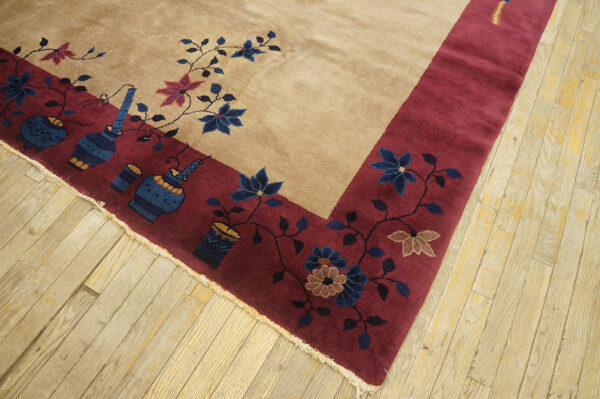 Tan field rug with a wide burgundy border featuring blue flowers and vase motifs, lying on light wood flooring.