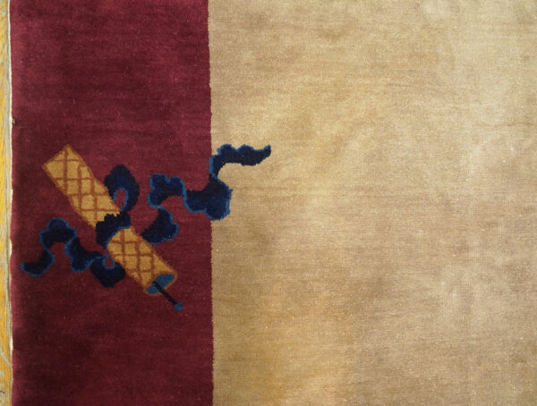 Maroon rug field with gold checkered scrolls and navy ribbons bordered by solid tan pile.