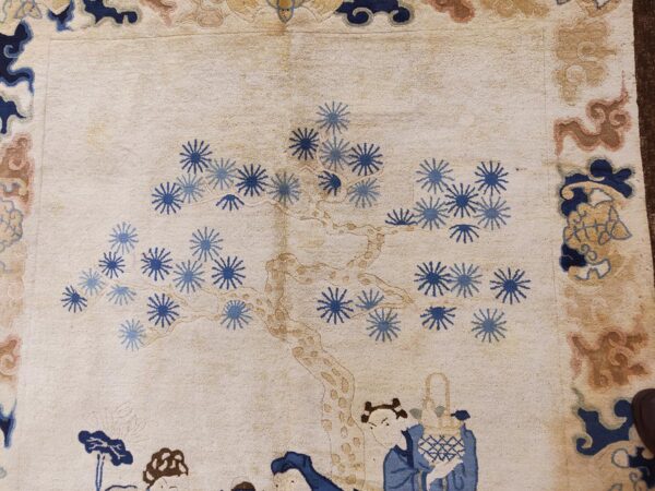 Cream rug with blue floral starbursts, a tan tree trunk, and figures along a blue and brown patterned border.