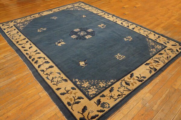 A large blue rug with a wide tan botanical border and scattered floral motifs rests on wood plank flooring.