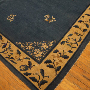 Low-pile blue rug with a tan border and stylized botanical motifs rests on light brown hardwood flooring.