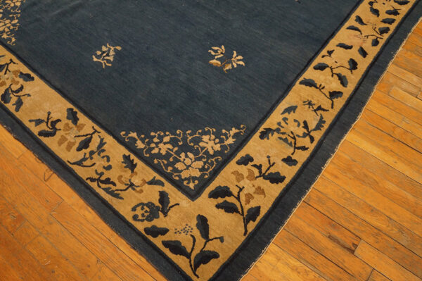 Low-pile blue rug with a tan border and stylized botanical motifs rests on light brown hardwood flooring.