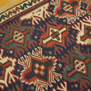 Flatweave rug featuring complex geometric motifs in navy, brick red, cream, and deep teal on wood planks.