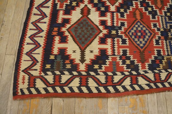 Geometric flat rug with red, cream, and navy stepped patterns and diamonds on light wooden flooring.
