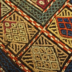 Intricate flatweave rug texture with red, blue, green, and gold geometric diamond patterns on dark ground.