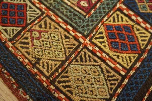 Intricate flatweave rug texture with red, blue, green, and gold geometric diamond patterns on dark ground.